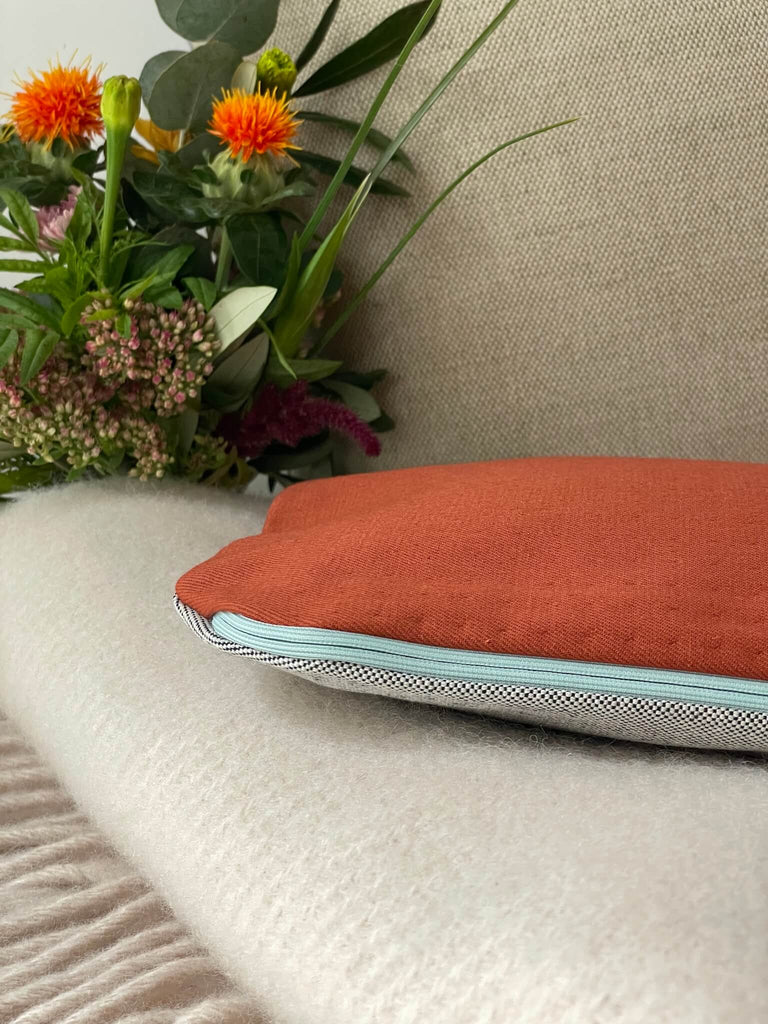 Detailansicht der Stoffe der handgefertigten Wärmflaschenhülle von HETTI., 100% Baumwolle in grau, orange mit hellblauem Reißverschluss, mit Blume und Kissen im Hintergrund